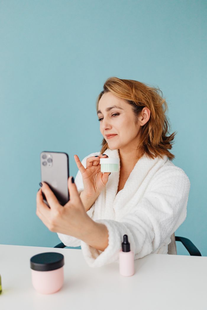 Smiling woman in robe taking selfie with skincare cream in modern setting.
